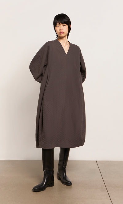 Plümo  Person wears a loose, dark brown Grenyer Dress from Plümo Studio and black knee-high boots against a plain background.