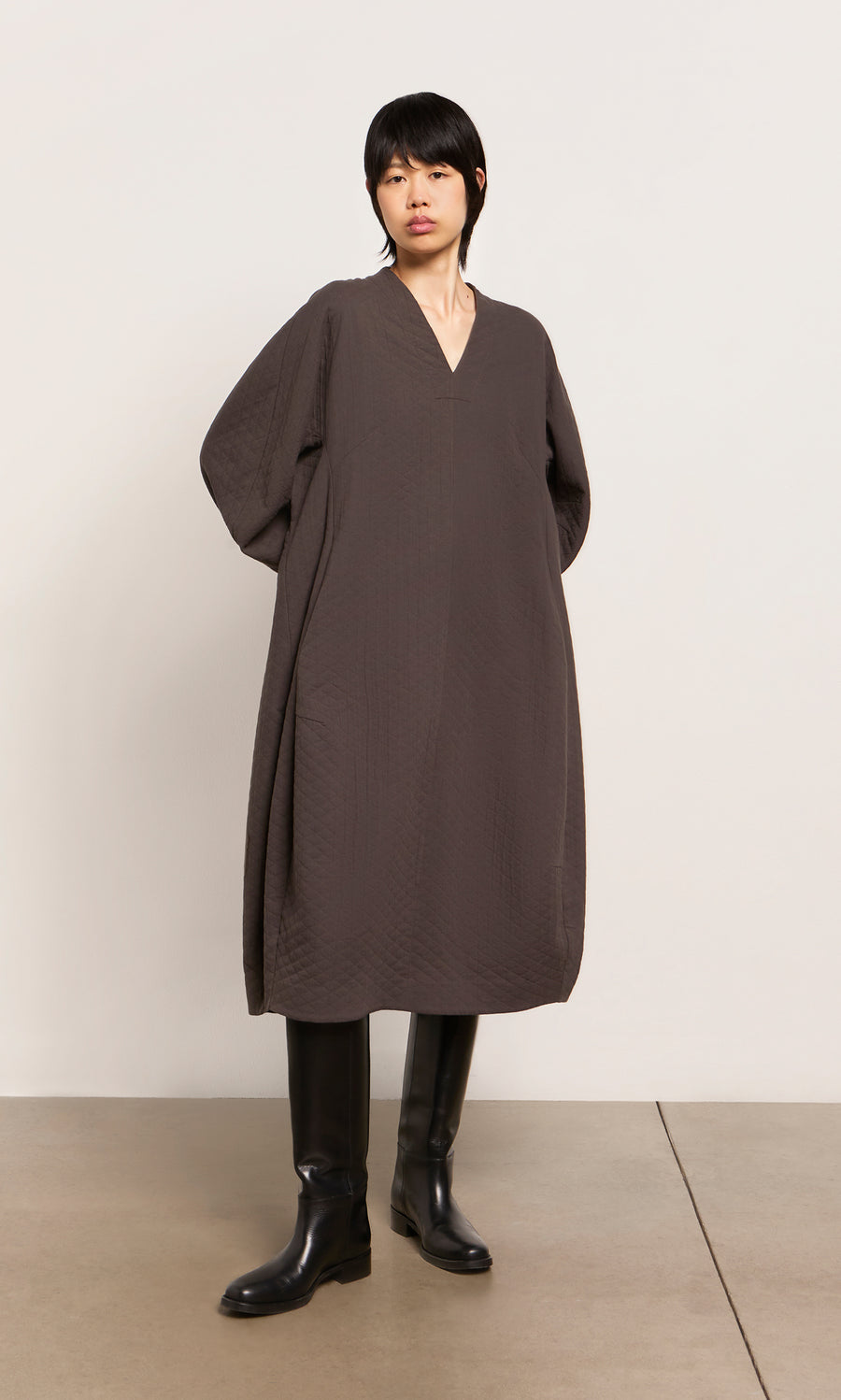 Plümo  Person wears a loose, dark brown Grenyer Dress from Plümo Studio and black knee-high boots against a plain background.