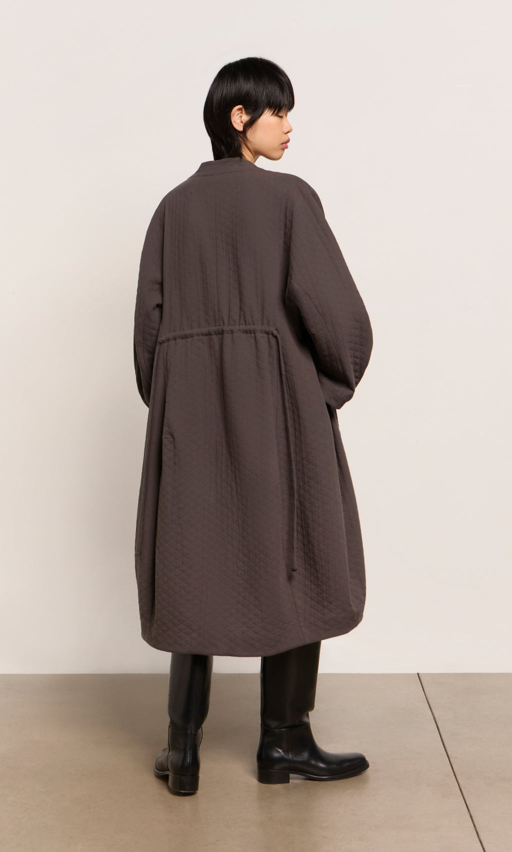 Plümo  Person with short dark hair wears the Grenyer Dress and a long brown coat, standing on a concrete floor.