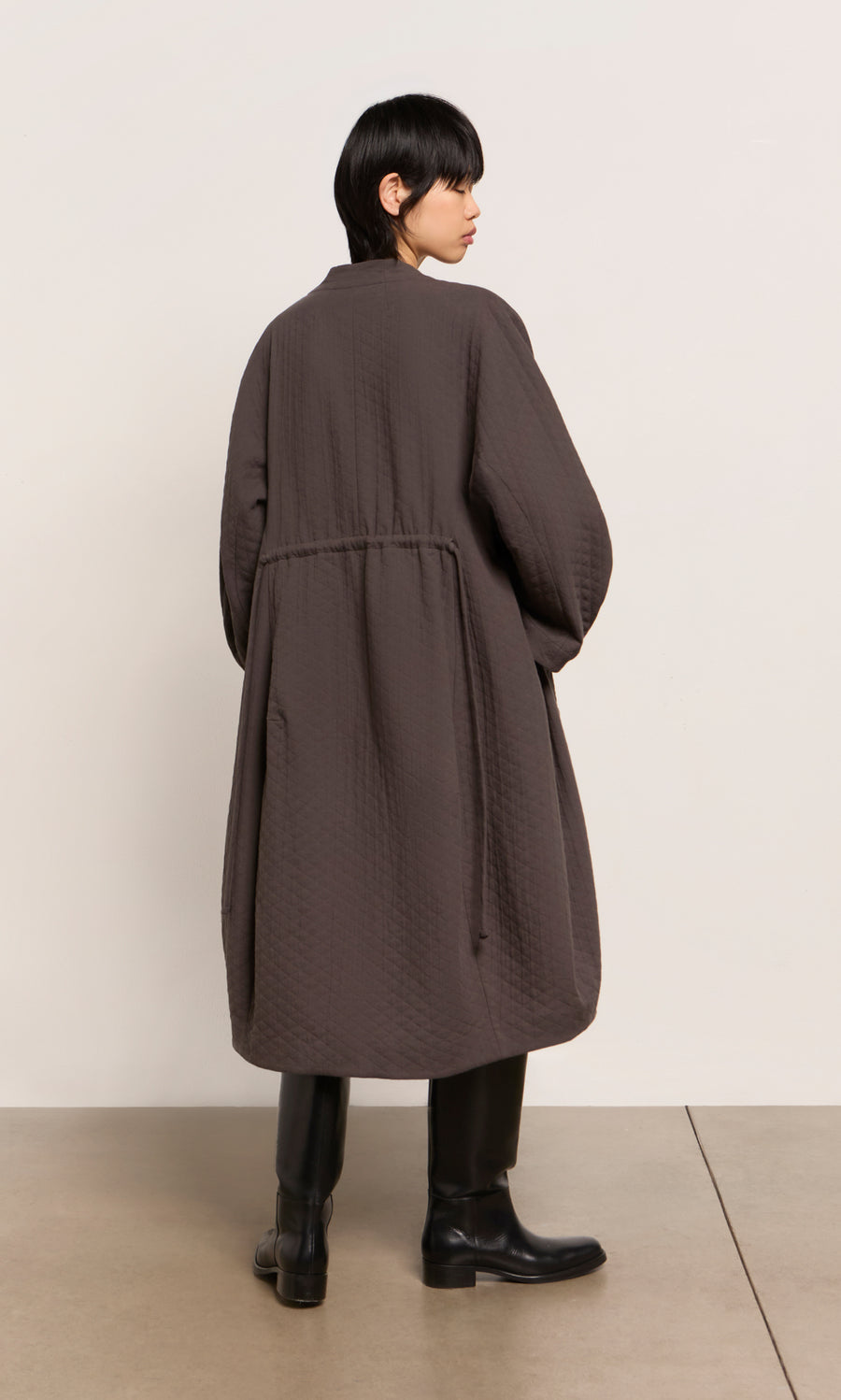 Plümo  Person with short dark hair wears the Grenyer Dress and a long brown coat, standing on a concrete floor.