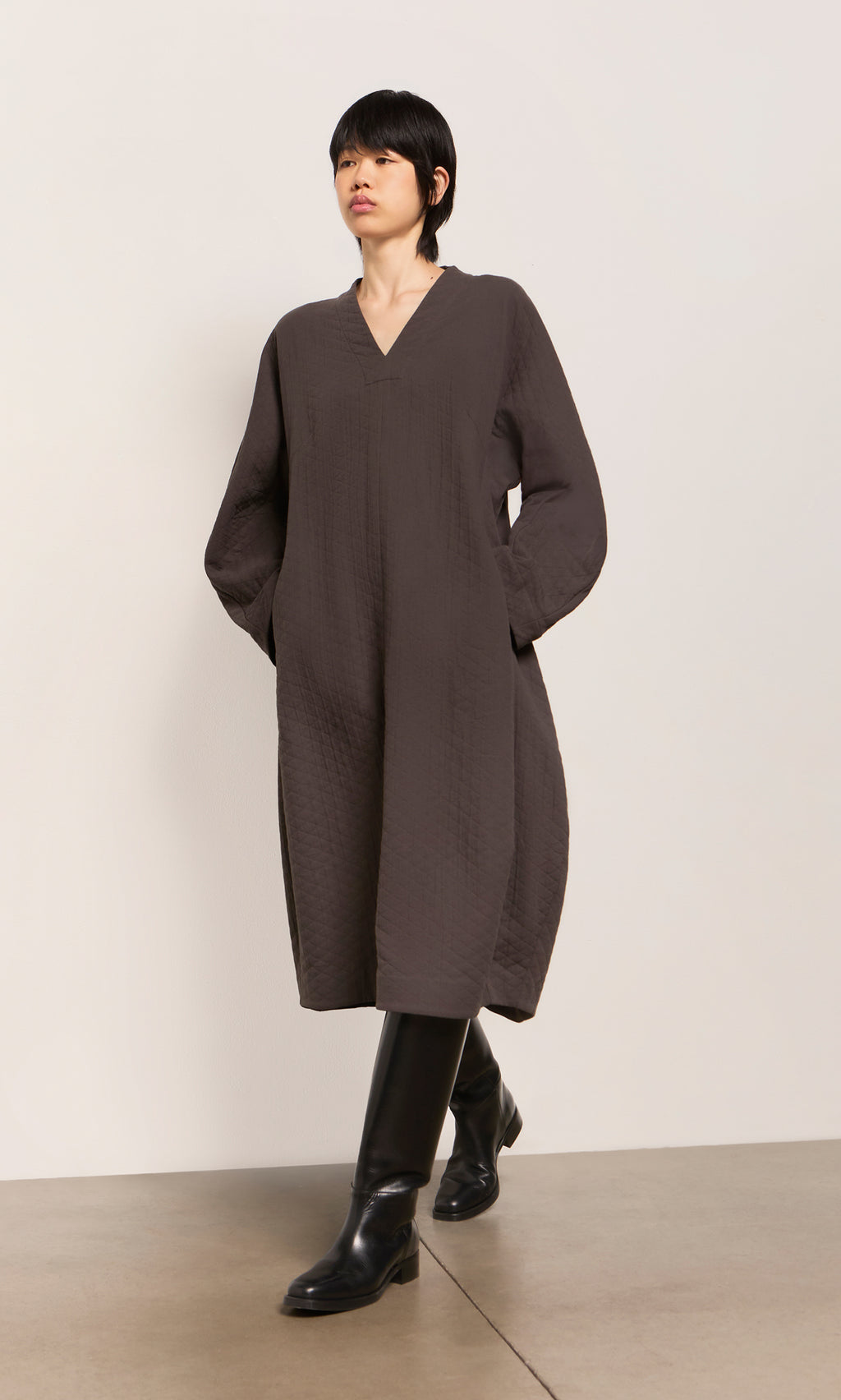 Plümo  Person in a loose, textured Grenyer Dress with long sleeves and tall black boots walking indoors.