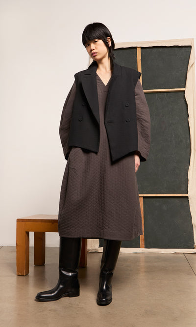 Plümo  Someone in a brown quilted dress, Volta Waistcoat, and boots stands indoors near a bench and abstract backdrop.