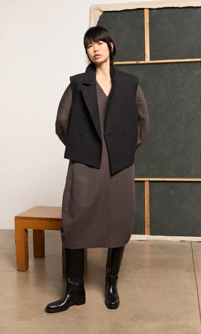 Plümo  Person in a long brown dress, Volta Waistcoat, and boots stands indoors with hands behind back.