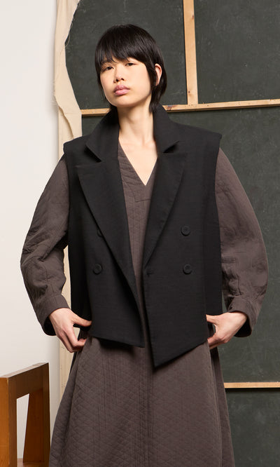 Plümo  Person in a textured brown dress and Volta Waistcoat stands before an art studio backdrop.