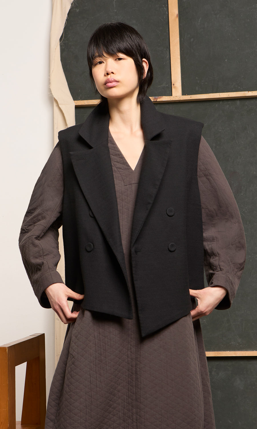 Plümo  Person in a textured brown dress and Volta Waistcoat stands before an art studio backdrop.