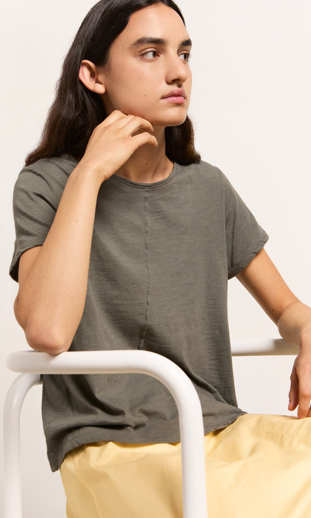 Plümo- Person with long dark hair in a Grey Cotton Tee and yellow pants sits on a white chair, looking to the side.