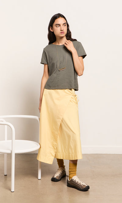 Plümo- Woman in a Grey Cotton Tee, yellow wrap skirt, and sneakers stands by a white chair indoors.
