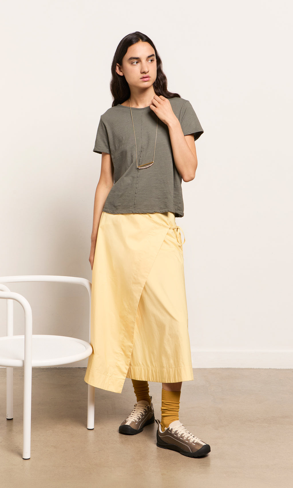 Plümo  Woman in gray tee, yellow wrap skirt, and breathable canvas Walking Shoes stands by a white chair indoors.