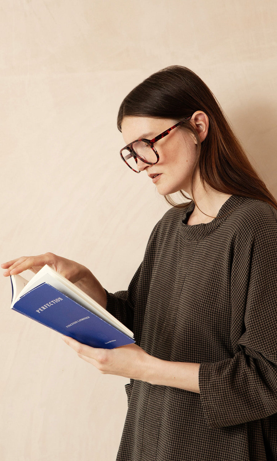 Plümo- Woman with long brown hair wears Havana Red Readers while reading a blue book against a neutral background.