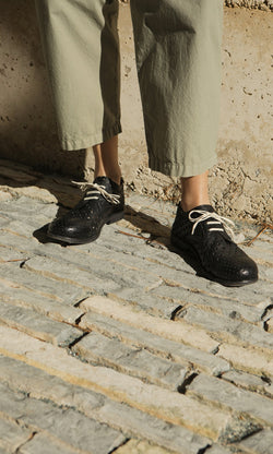 Plümo- Person wearing light green pants and Black Derby Brogues, standing on textured stone pavement in sunlight.