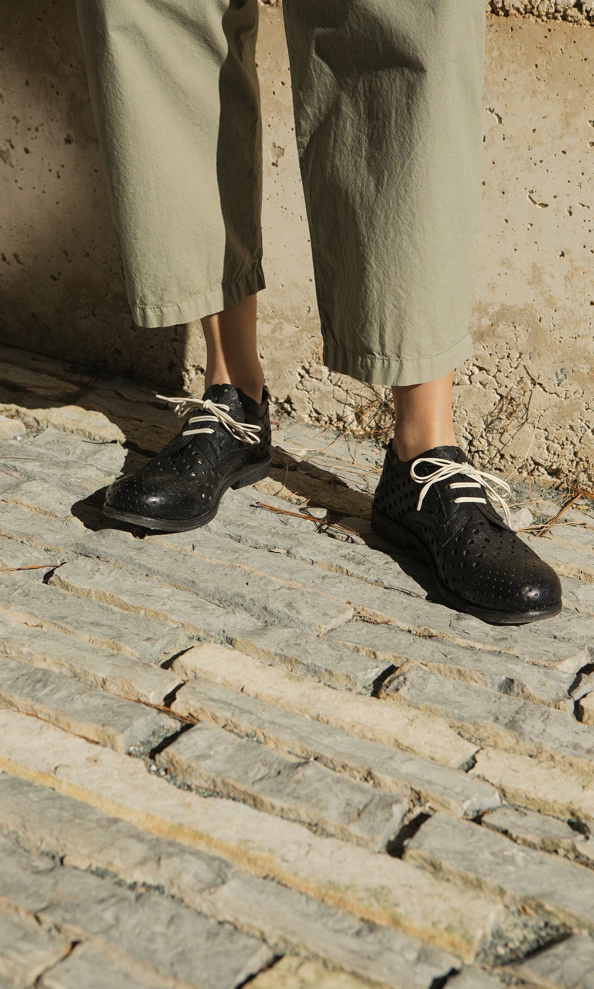 Plümo- Person wearing light green pants and Black Derby Brogues, standing on textured stone pavement in sunlight.
