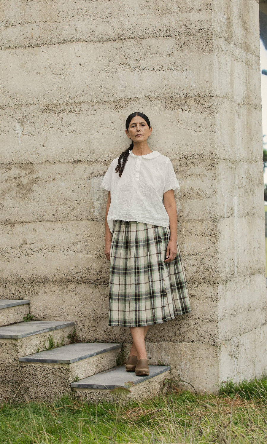 Plümo- A woman in a white blouse and the Hawksmoor Kilt stands on outdoor steps by a tall wall, looking ahead.