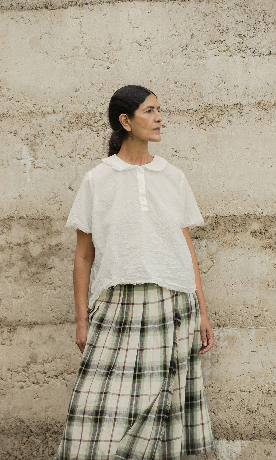 Plümo- Woman in the Renville Shirt stands against a textured beige wall, looking to the side.