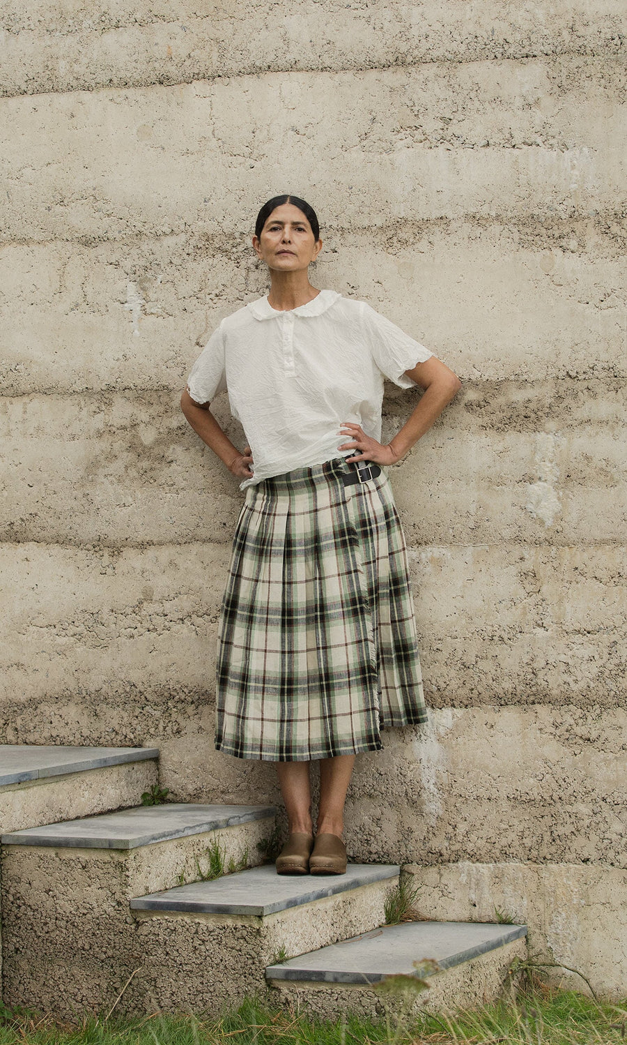 Plümo- A person stands on stone steps in Dublin by a concrete wall, wearing a white shirt and the Hawksmoor Kilt.