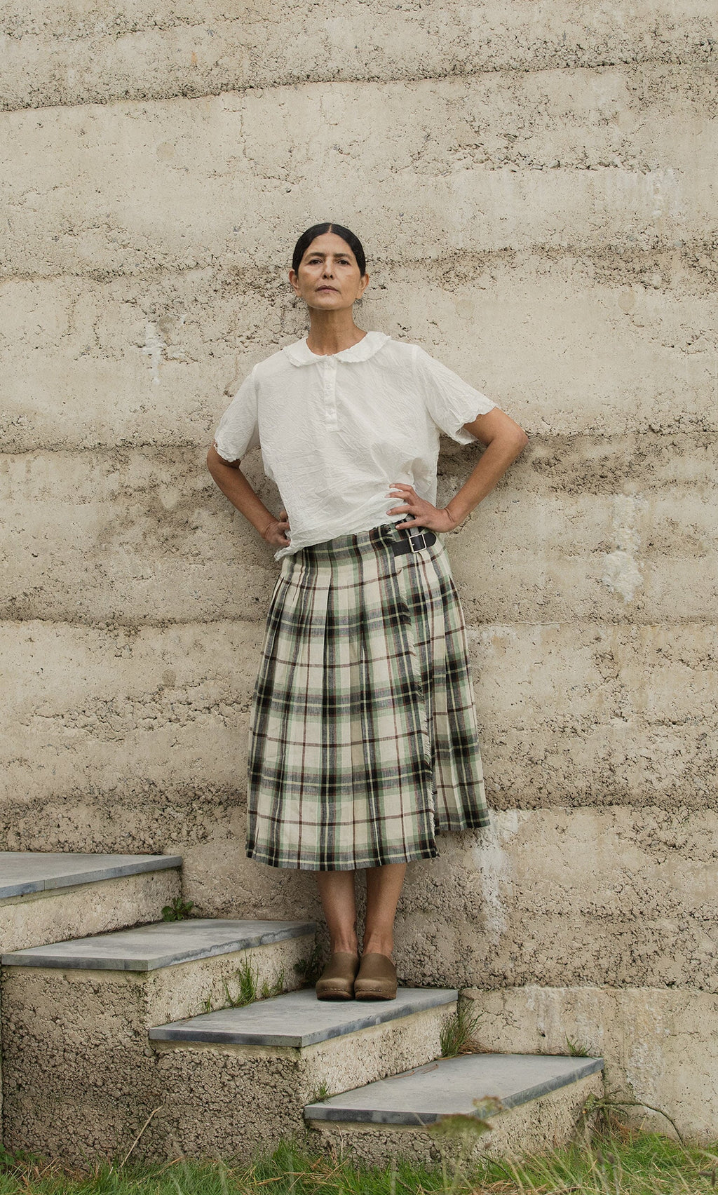 Plümo- A woman stands on stone steps, wearing the white cotton Renville Shirt and a plaid skirt by a textured beige wall.