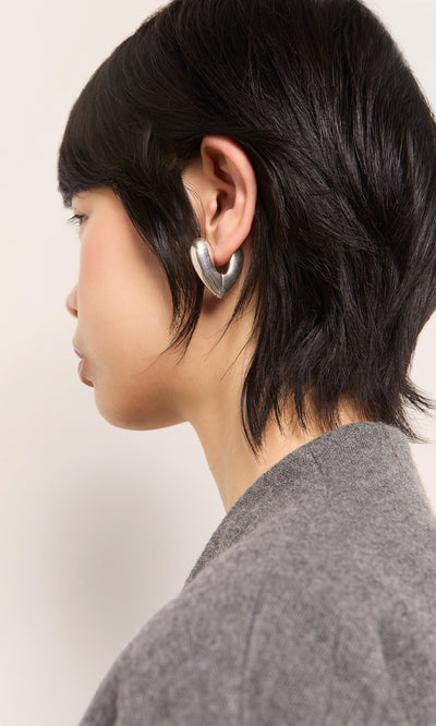 Plümo  Person with short dark hair in profile wears Silver Heart Earrings and a gray textured top.