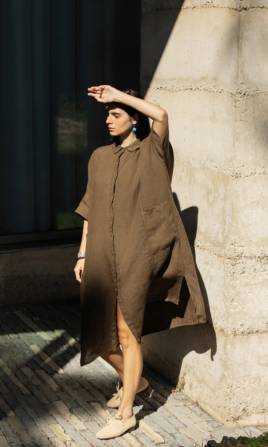 Plümo- Wearing the Hockley Linen Dress, a person stands in sunlight, shielding their eyes near a concrete wall.