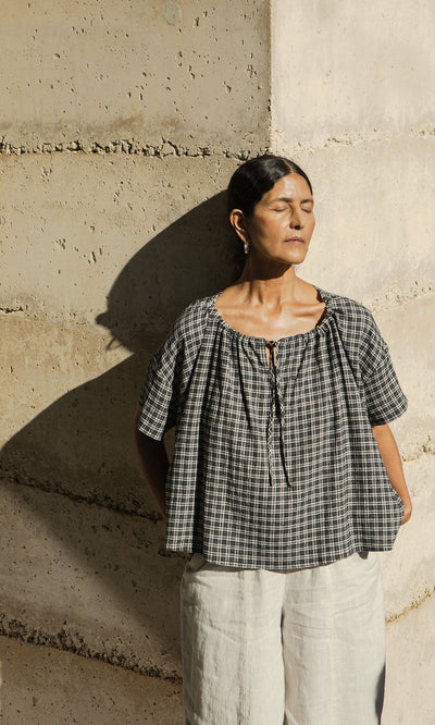 Plümo- A woman in the Ida Tie Blouse - Plaid and light pants stands against a concrete wall, eyes closed, enjoying sunlight.