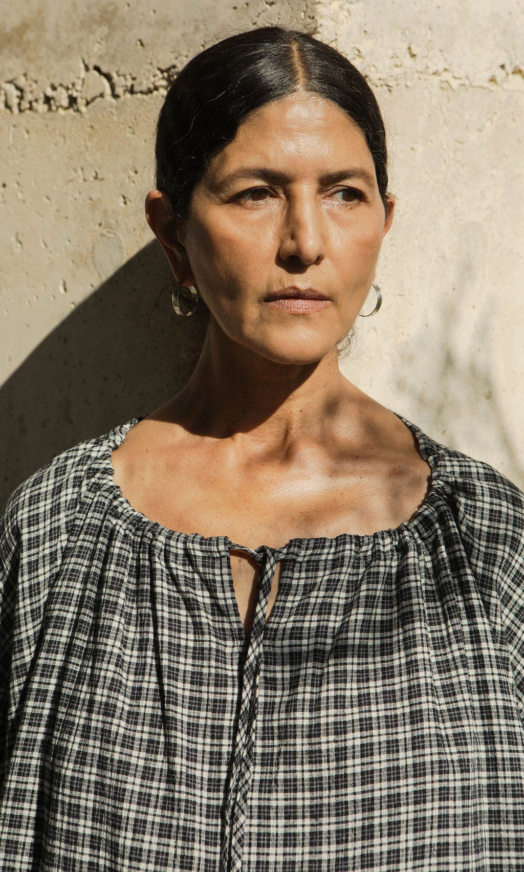 Plümo- A woman with dark hair pulled back wears Hui Hoops and a plaid top, standing by a sunlit textured wall.
