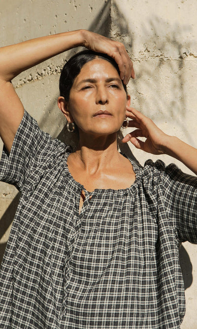 Plümo- Woman in the Ida Tie Blouse - Plaid stands in sunlight with one arm raised, eyes closed, against a textured wall.