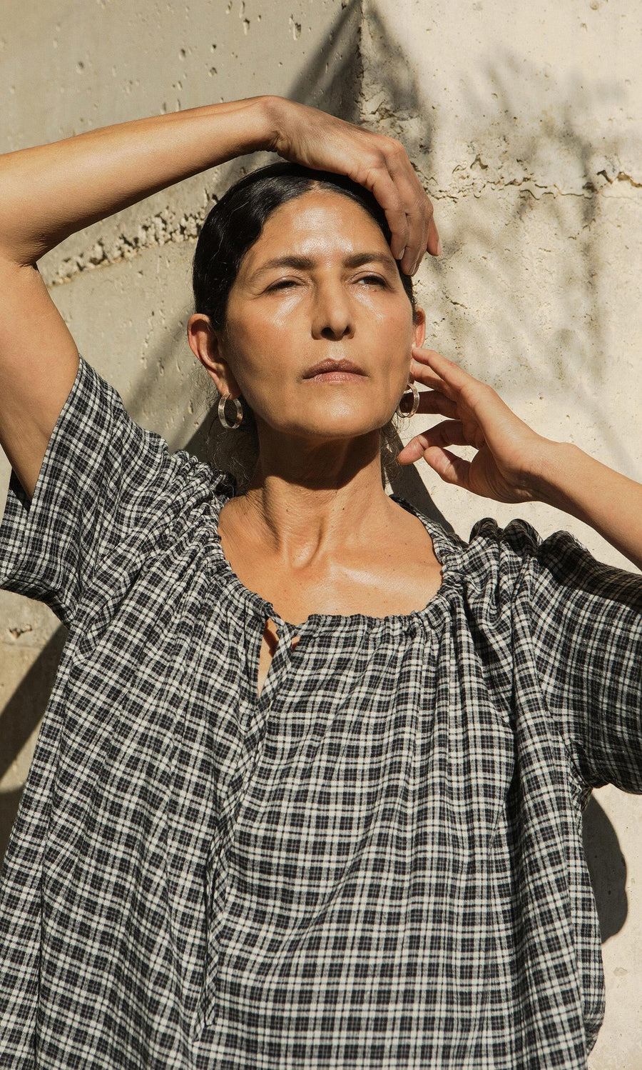 Plümo- Woman in the Ida Tie Blouse - Plaid stands in sunlight with one arm raised, eyes closed, against a textured wall.