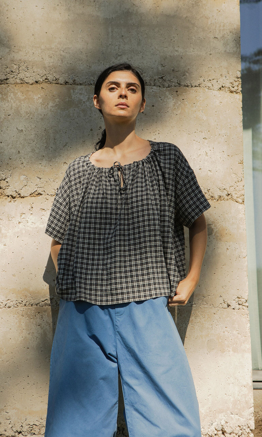 Plümo- Woman in the Ida Tie Blouse - Plaid stands against a stone wall, gazing upward with confidence.