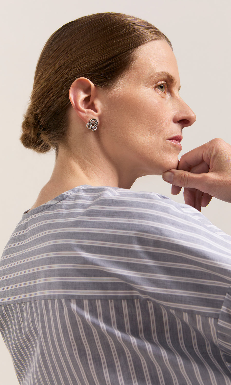 Plümo  Woman in striped shirt, low bun, wears Infinity Earrings and looks thoughtful with her hand on her chin.