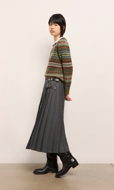 Plümo  Person in a patterned sweater, Boyd Kilt in grey worsted wool, and black boots standing sideways by a plain wall.