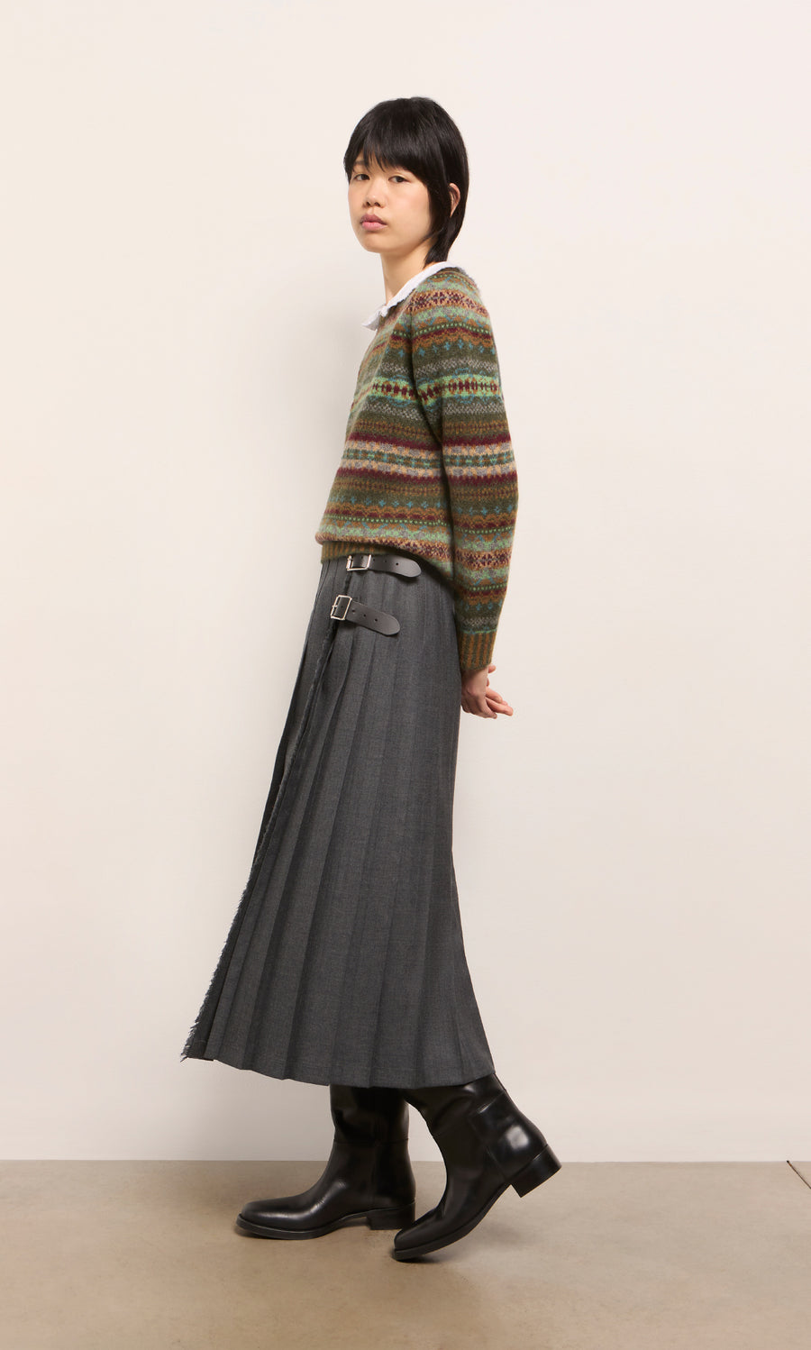 Plümo  Person in a patterned sweater, Boyd Kilt in grey worsted wool, and black boots standing sideways by a plain wall.