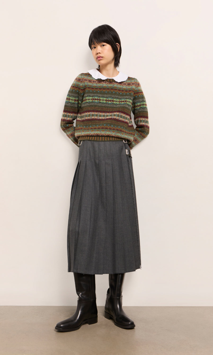 Plümo  Woman in the Iona Fairisle Jumper, white shirt, gray pleated skirt, and black boots stands by a plain wall.