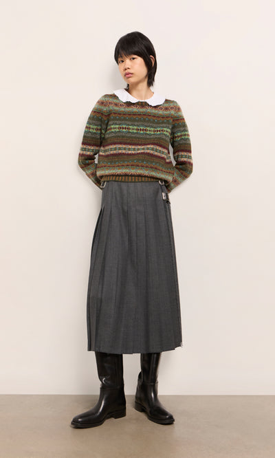 Plümo  Woman in a patterned sweater, Boyd Kilt, white collared shirt, and black boots stands against a plain wall.