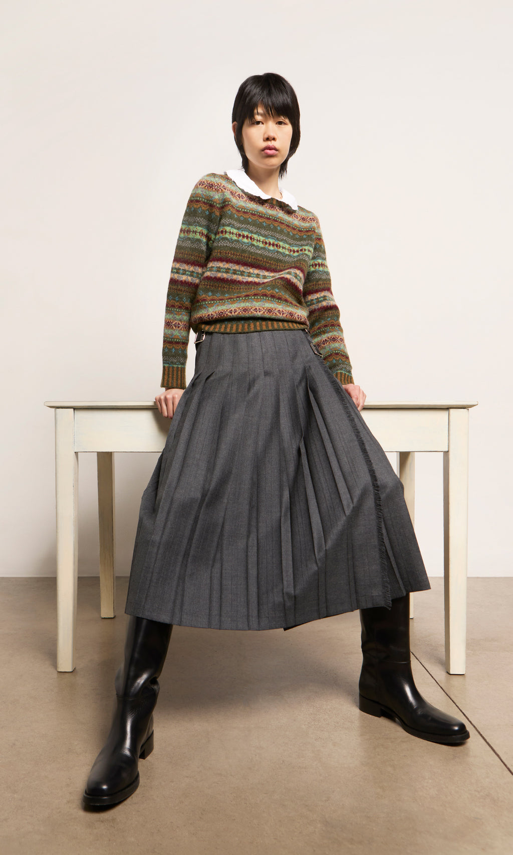 Plümo  Person in an Iona Fairisle Jumper, pleated gray skirt, and black boots sits on a white desk with legs apart.