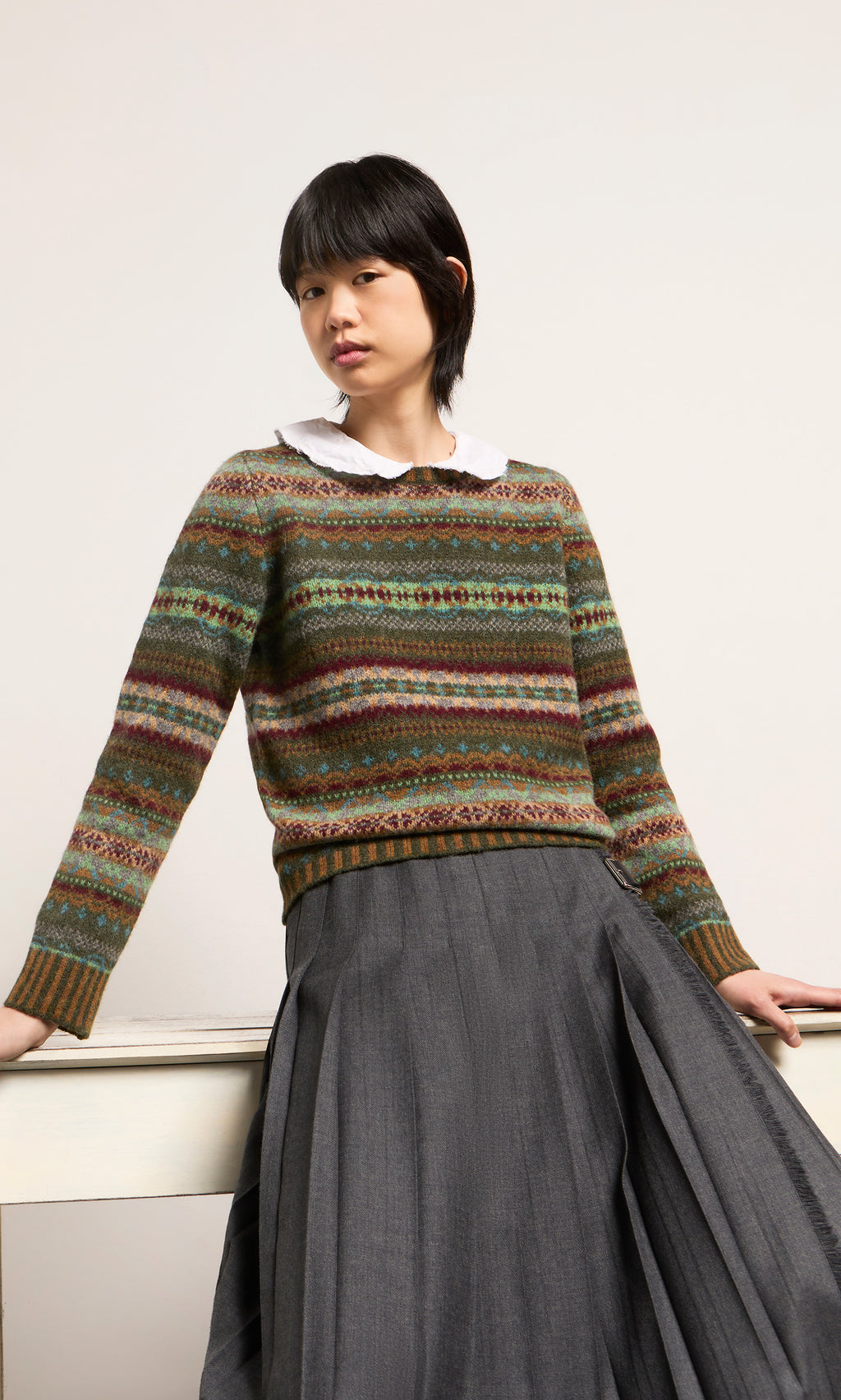Plümo  Person in an Iona Fairisle Jumper and gray pleated skirt leans on a white table against a plain background.