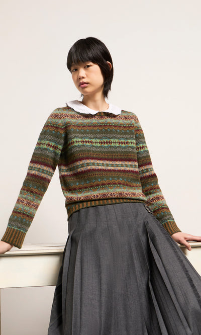 Plümo  Person in an Iona Fairisle Jumper and gray pleated skirt leans on a white table against a plain background.