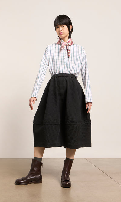 Plümo  Person stands in a striped shirt, black midi skirt, brown boots, and a Match Necktie against a plain background.