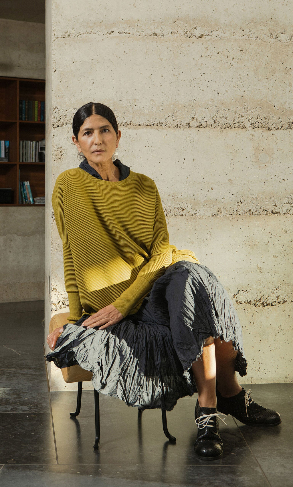 Plümo- Woman in a Jack Sweater - Burnt Gold by Plümo Studio sits on a chair, modern room and bookshelves behind her.