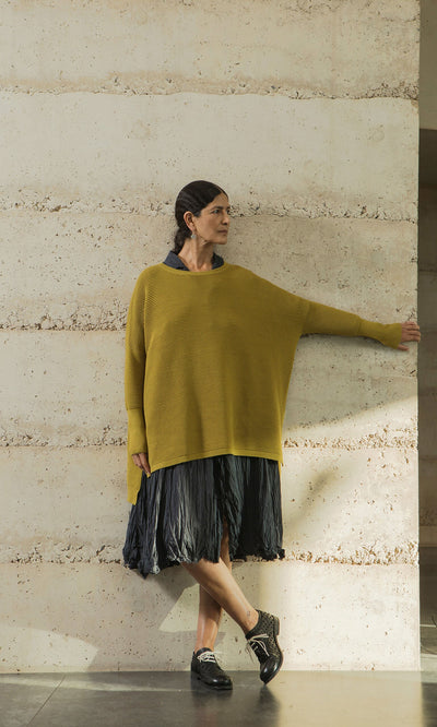 Plümo- Woman in the Jack Sweater - Burnt Gold and a black skirt leans against a textured wall, looking to her left.