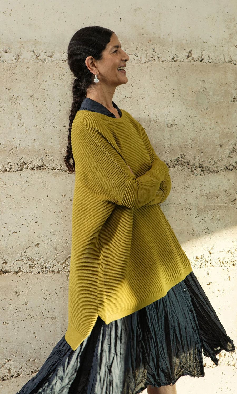 Plümo- Woman with dark hair in a braid wears the Jack Sweater - Burnt Gold by Plümo Studio, smiling by a textured wall.