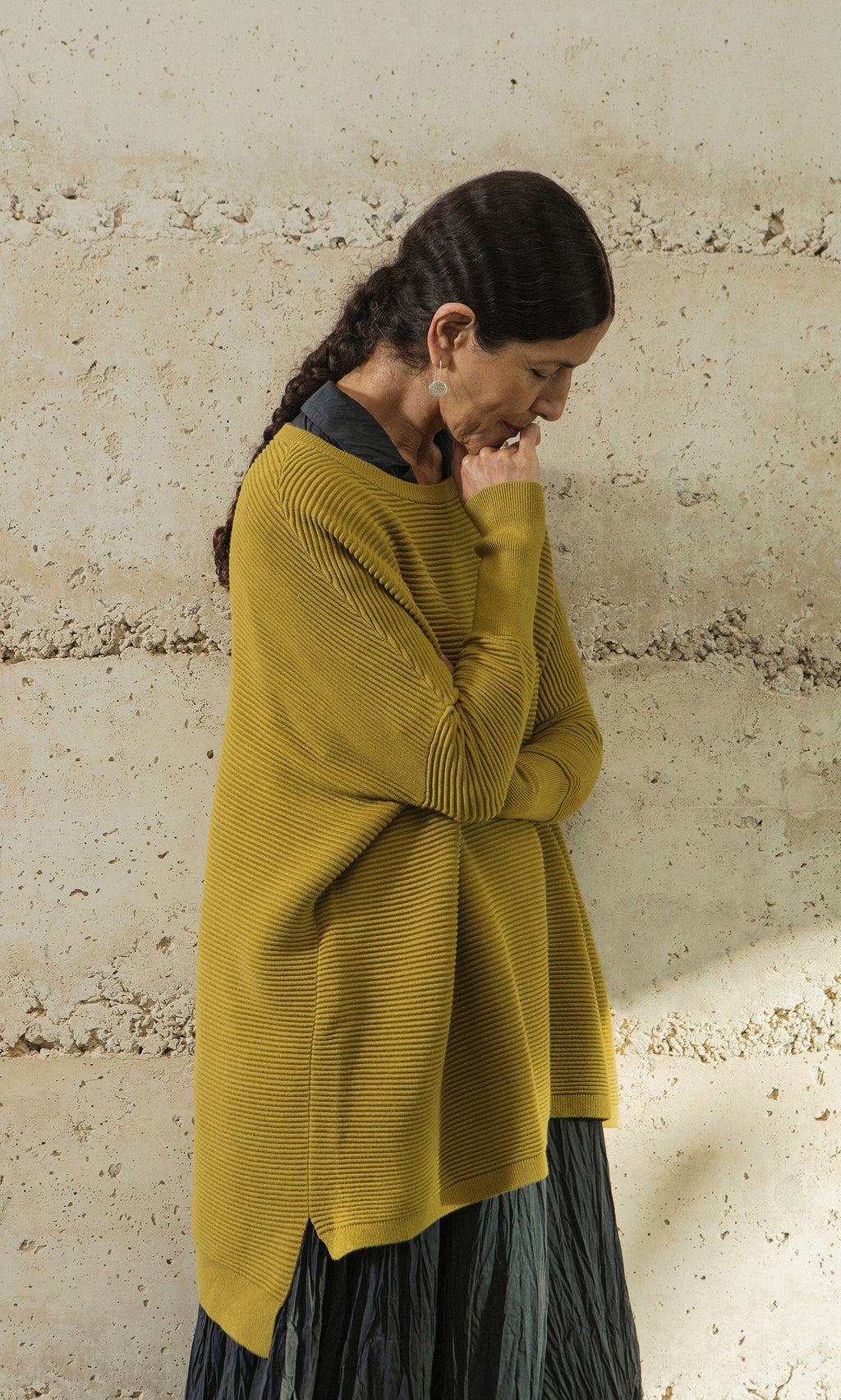 Plümo- A woman in the Jack Sweater - Burnt Gold stands against a textured wall, looking down with her hand on her chin.