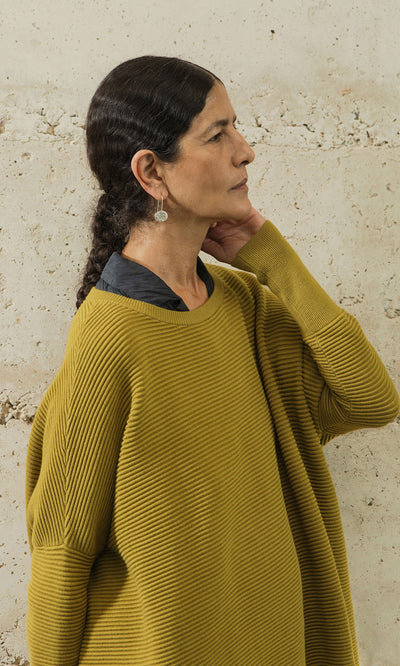 Plümo- Woman with dark hair in a braid wears the Jack Sweater in Burnt Gold, standing sideways and touching her neck.