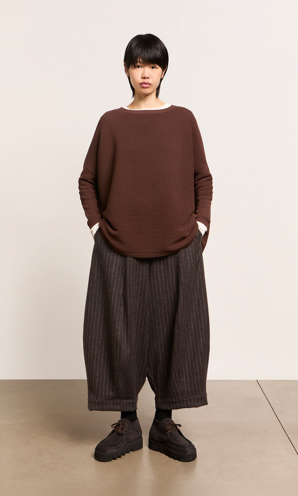 Plümo  Person stands facing forward in Rolla Boots, loose brown sweater, striped pants; hands in pockets.
