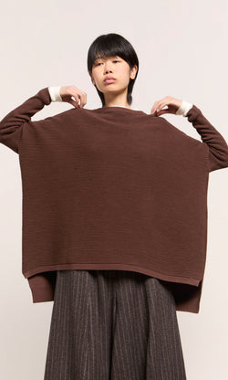 Plümo  Person wearing the Jack Sweater - Bitter Chocolate, holding it up to show its back, paired with striped pants.
