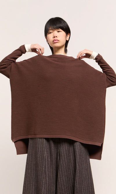 Plümo  Person wearing the Jack Sweater - Bitter Chocolate, holding it up to show its back, paired with striped pants.