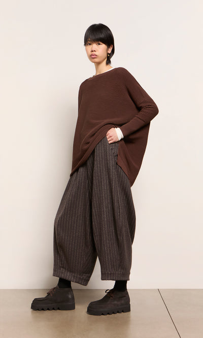 Plümo  Person in Jack Sweater - Bitter Chocolate, wide pinstriped pants, and platforms stands against a light wall.