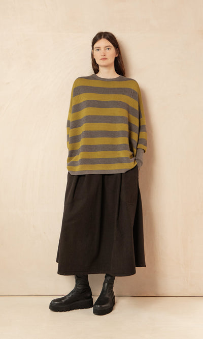 Plümo- A woman stands against a beige wall in a Jack Sweater - Butter Stripe, black skirt, and black boots.