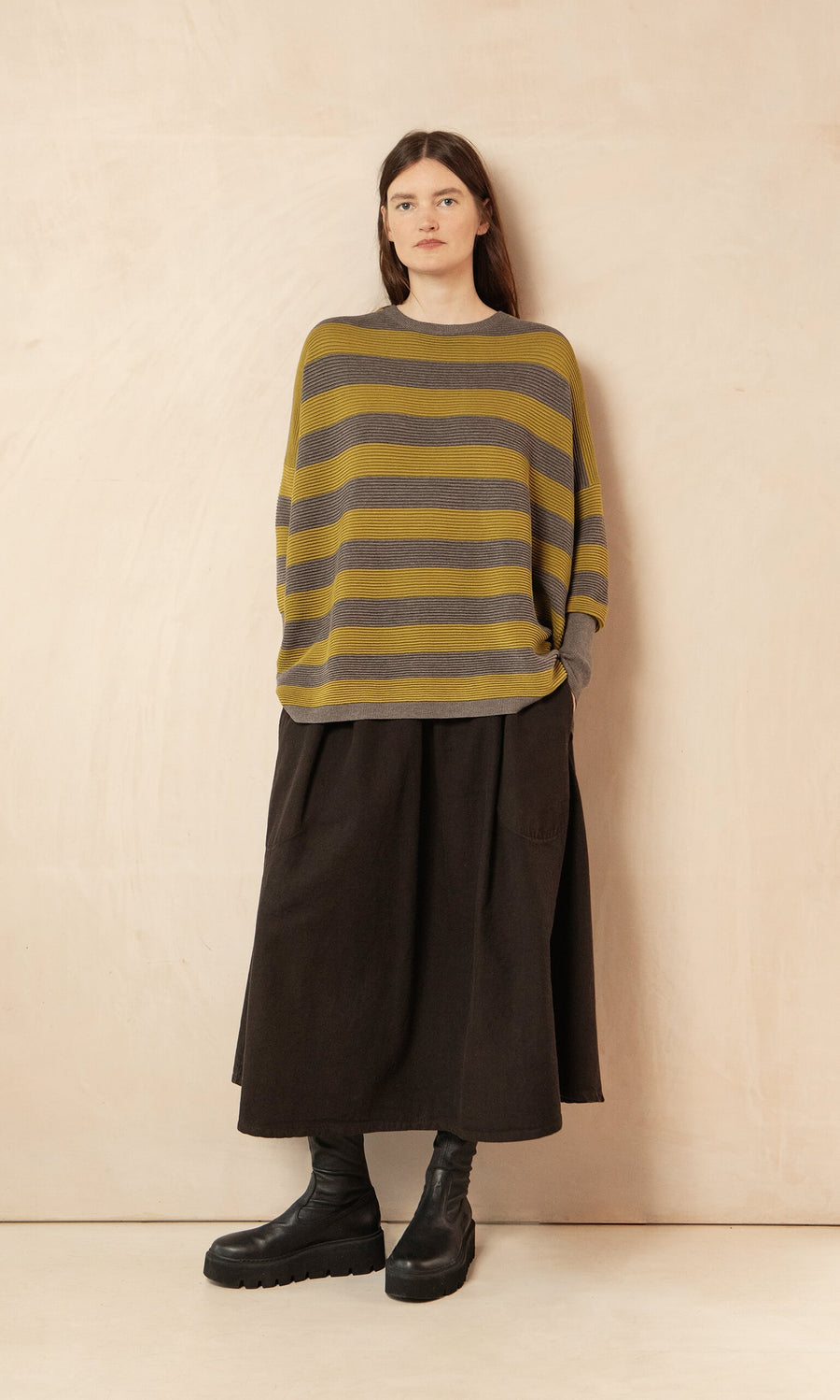 Plümo- A woman stands against a beige wall in a Jack Sweater - Butter Stripe, black skirt, and black boots.