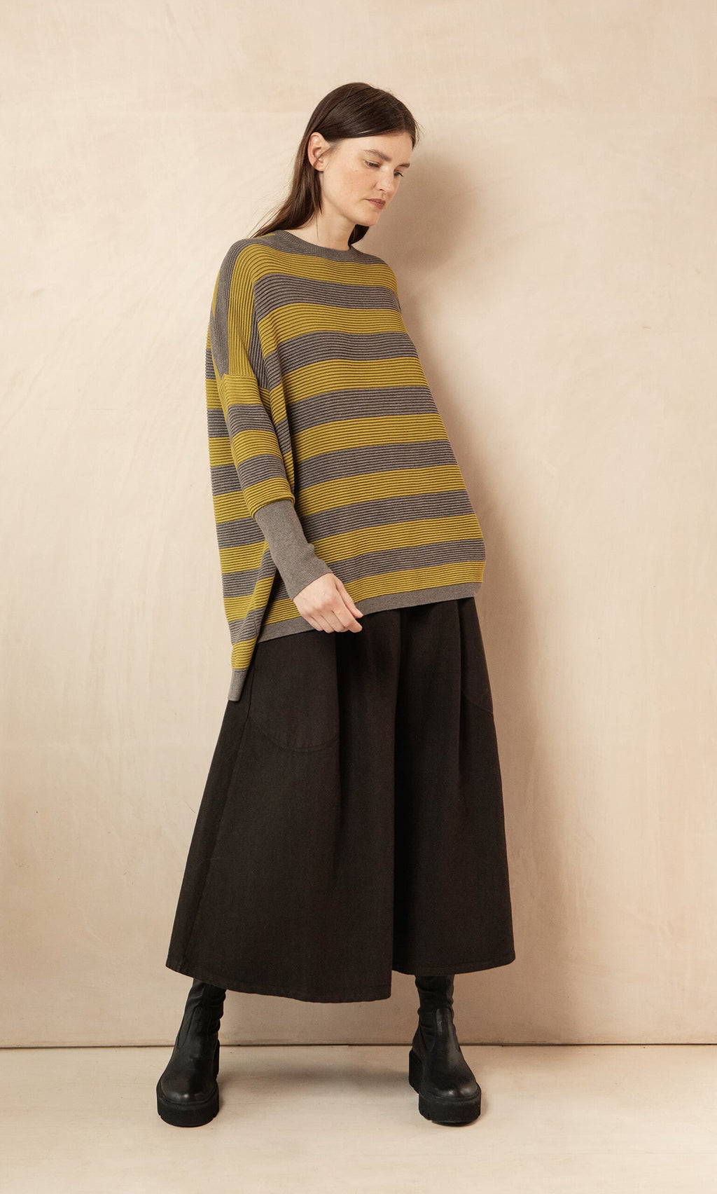 Plümo- Woman in the Jack Sweater - Butter Stripe by Plümo Studio, wide black pants, and boots stands against a beige wall.