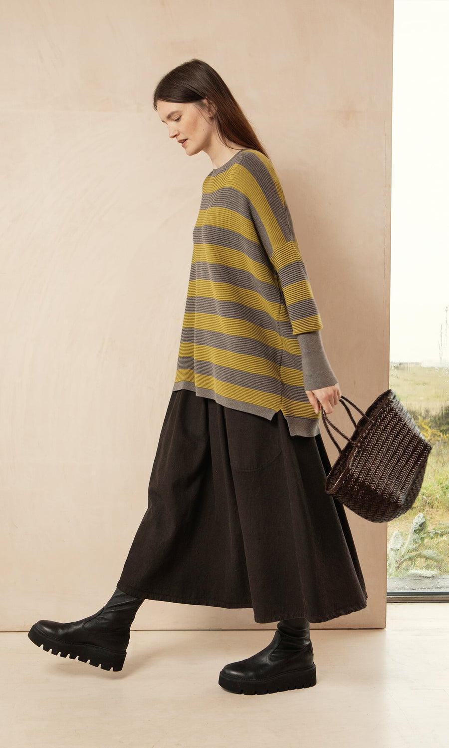 Plümo- Woman in the Jack Sweater - Butter Stripe by Plümo Studio, long black skirt, boots, holds a dark woven handbag.