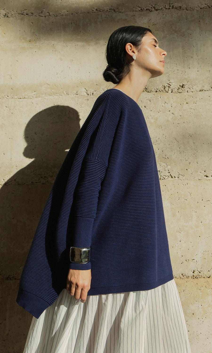 Plümo- Woman wearing the Jack Sweater - Navy with dropped shoulders and a white skirt stands sideways in sunlight.