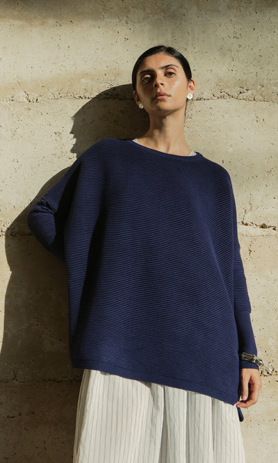 Plümo- Person in the Jack Sweater - Navy and a white pleated skirt stands by a light stone wall in sunlight.